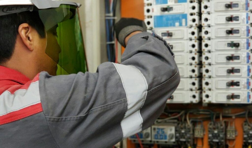 An electrician in a hard hat and face shield testing an electrical panel